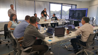 Participants sitting in a u-shape with laptops, and large screen showing demo.